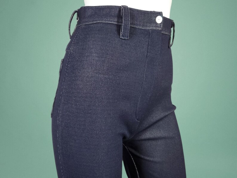 May include: Close-up of dark blue denim trousers. The trousers feature a high waist, belt loops, and a button closure. The fabric has a subtle texture, and the stitching is visible. These are a classic style, suitable for various outfits.