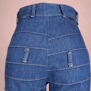 K&ouml;nnte beinhalten: Nahaufnahme einer blauen Jeanshose mit einem einzigartigen horizontalen Paneeldesign. Die Hose hat zwei Ges&auml;&szlig;taschen und G&uuml;rtelschlaufen. Der Denim hat eine mittlere Waschung und sichtbare Nahtdetails.