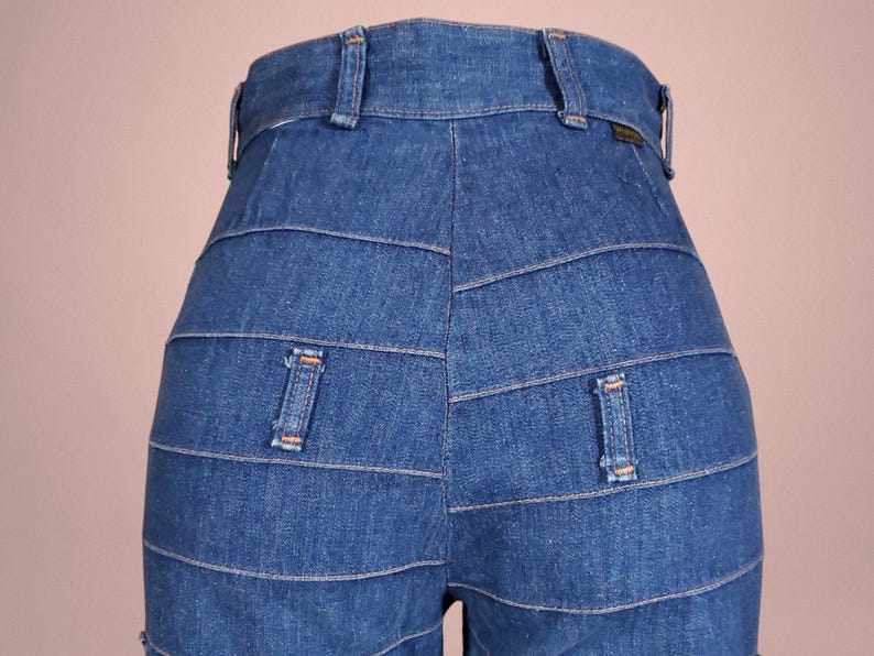 K&ouml;nnte beinhalten: Nahaufnahme einer blauen Jeanshose mit einem einzigartigen horizontalen Paneeldesign. Die Hose hat zwei Ges&auml;&szlig;taschen und G&uuml;rtelschlaufen. Der Denim hat eine mittlere Waschung und sichtbare Nahtdetails.