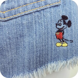 K&ouml;nnte beinhalten: Eine blaue Jeans-Shorts mit ausgefranstem Saum und einem gestickten Mickey-Mouse-Motiv.