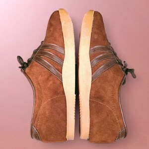 May include: A pair of brown suede sneakers with white soles and brown laces. The sneakers have a classic design with three brown stripes on each side.