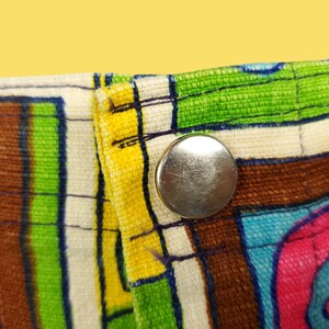 Peut inclure: Gros plan sur une section de tissu avec un motif abstrait color&eacute;. Le motif pr&eacute;sente des rayures et des formes audacieuses en rose, vert, bleu, marron et jaune. Un bouton-pression en m&eacute;tal argent&eacute; est visible sur le bord du tissu.