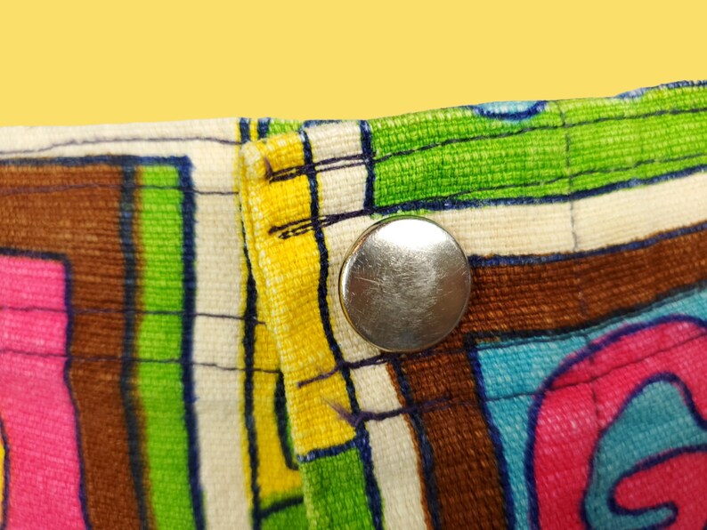 Peut inclure: Gros plan sur une section de tissu avec un motif abstrait color&eacute;. Le motif pr&eacute;sente des rayures et des formes audacieuses en rose, vert, bleu, marron et jaune. Un bouton-pression en m&eacute;tal argent&eacute; est visible sur le bord du tissu.