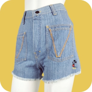 K&ouml;nnte beinhalten: Hellblaue Jeans-Shorts mit ausgefransten Kanten und einem gestickten Mickey Mouse-Design in Gelb auf der Ges&auml;&szlig;tasche.