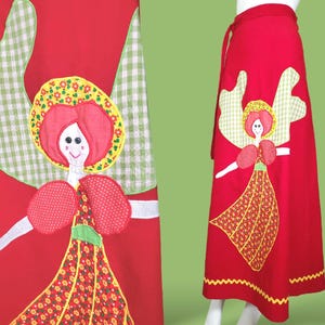May include: A red wrap skirt with a patchwork design featuring a smiling angel with green wings, a yellow hat, and a red and yellow dress. The skirt has a scalloped hem.