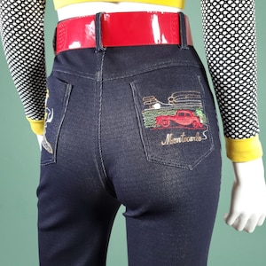May include: Dark blue denim jeans with a red patent leather belt. The back pocket features an embroidered design of a red car and the words "Montecarlo S". The model is wearing a yellow top and a black and white polka dot sleeve.