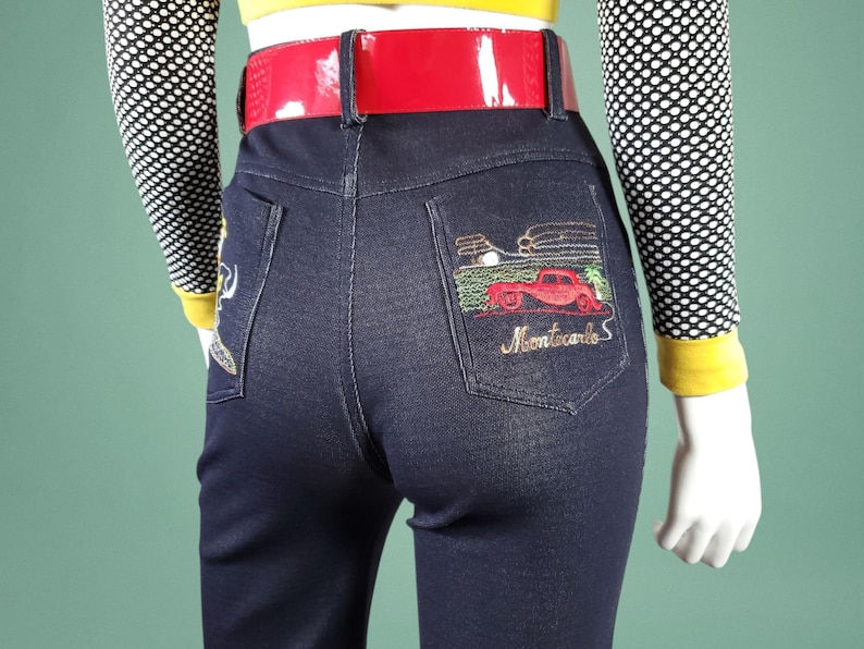 May include: Dark blue denim jeans with a red patent leather belt. The back pocket features an embroidered design of a red car and the words "Montecarlo S". The model is wearing a yellow top and a black and white polka dot sleeve.