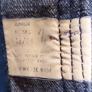 K&ouml;nnte beinhalten: Nahaufnahme eines Stoffetiketts, das auf Denim gen&auml;ht ist. Das Etikett ist beige mit schwarzem Text, der "JUNIOR", "MOSES" und "13/14" lautet. Zus&auml;tzlicher Text enth&auml;lt Pflegehinweise. Das Etikett ist mit dunkelblauem Faden gen&auml;ht.