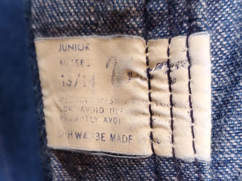 K&ouml;nnte beinhalten: Nahaufnahme eines Stoffetiketts, das auf Denim gen&auml;ht ist. Das Etikett ist beige mit schwarzem Text, der "JUNIOR", "MOSES" und "13/14" lautet. Zus&auml;tzlicher Text enth&auml;lt Pflegehinweise. Das Etikett ist mit dunkelblauem Faden gen&auml;ht.