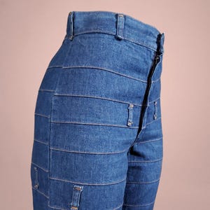 K&ouml;nnte beinhalten: Nahaufnahme einer blauen Jeans mit einem einzigartigen horizontalen Panel-Design. Die Jeans hat einen Rei&szlig;verschluss und G&uuml;rtelschlaufen. Das Kleidungsst&uuml;ck ist aus einem mittelblauen Denim-Stoff gefertigt.