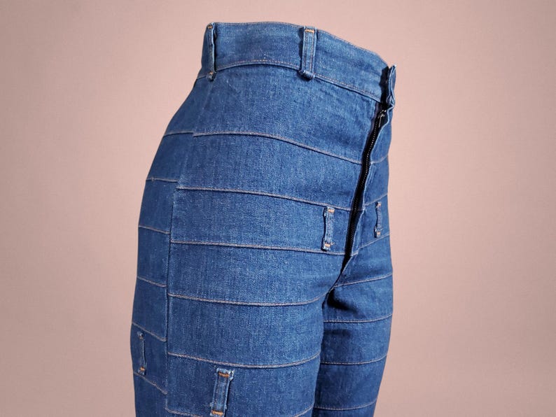 K&ouml;nnte beinhalten: Nahaufnahme einer blauen Jeans mit einem einzigartigen horizontalen Panel-Design. Die Jeans hat einen Rei&szlig;verschluss und G&uuml;rtelschlaufen. Das Kleidungsst&uuml;ck ist aus einem mittelblauen Denim-Stoff gefertigt.