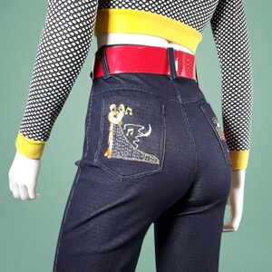 May include: Dark blue denim jeans with embroidered musical instrument design on the back pockets. The jeans have a red belt and are paired with a black and white polka dot top with yellow trim.