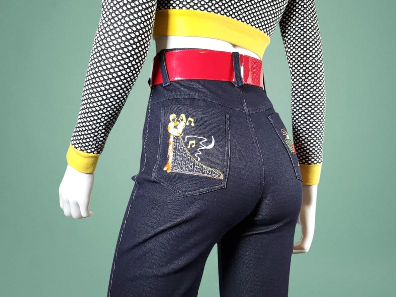 May include: Dark blue denim jeans with embroidered musical instrument design on the back pockets. The jeans have a red belt and are paired with a black and white polka dot top with yellow trim.