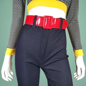 May include: A mannequin displays a vintage ensemble. It includes dark blue, high-waisted trousers, a glossy red belt, and a black and white polka dot top with yellow trim. The outfit is a striking fashion choice.