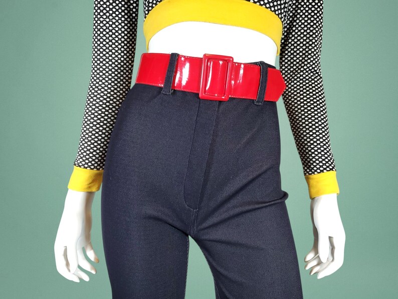 May include: A mannequin displays a vintage ensemble. It includes dark blue, high-waisted trousers, a glossy red belt, and a black and white polka dot top with yellow trim. The outfit is a striking fashion choice.
