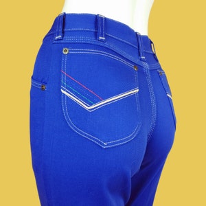 May include: Blue denim jeans with a white stitched back pocket and colourful stitching detail.