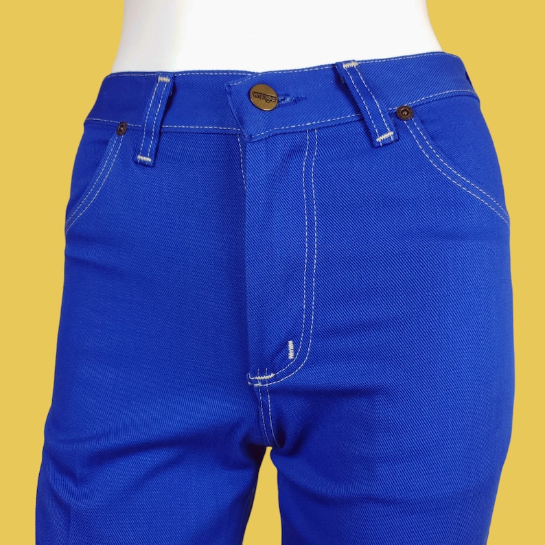 May include: A pair of blue denim jeans with white stitching. The jeans have a button fly and a small brown leather patch with the word "Wrangler" on the back.