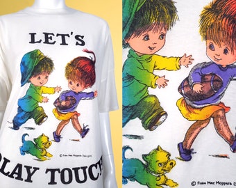 Peut inclure: T-shirt blanc avec un motif arc-en-ciel de deux enfants jouant au football américain avec un chien. Le texte "Let's Play Touch" est imprimé sur le t-shirt.