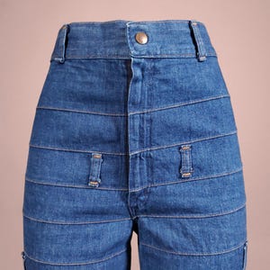 K&ouml;nnte beinhalten: Nahaufnahme einer blauen Jeanshose mit hohem Bund und Knopfverschluss. Die Hose hat horizontale, gen&auml;hte Bahnen auf der Vorderseite und kleine dekorative Taschen. Der Denim hat eine mittlere Waschung.