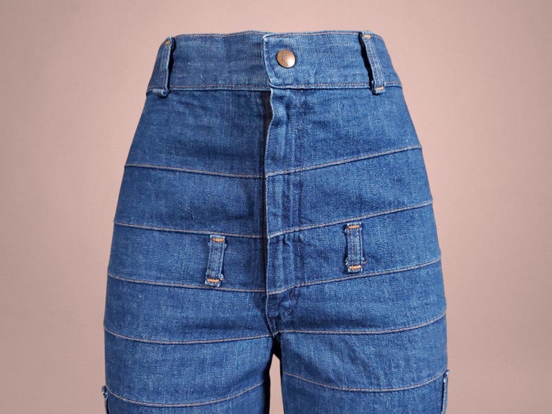 K&ouml;nnte beinhalten: Nahaufnahme einer blauen Jeanshose mit hohem Bund und Knopfverschluss. Die Hose hat horizontale, gen&auml;hte Bahnen auf der Vorderseite und kleine dekorative Taschen. Der Denim hat eine mittlere Waschung.