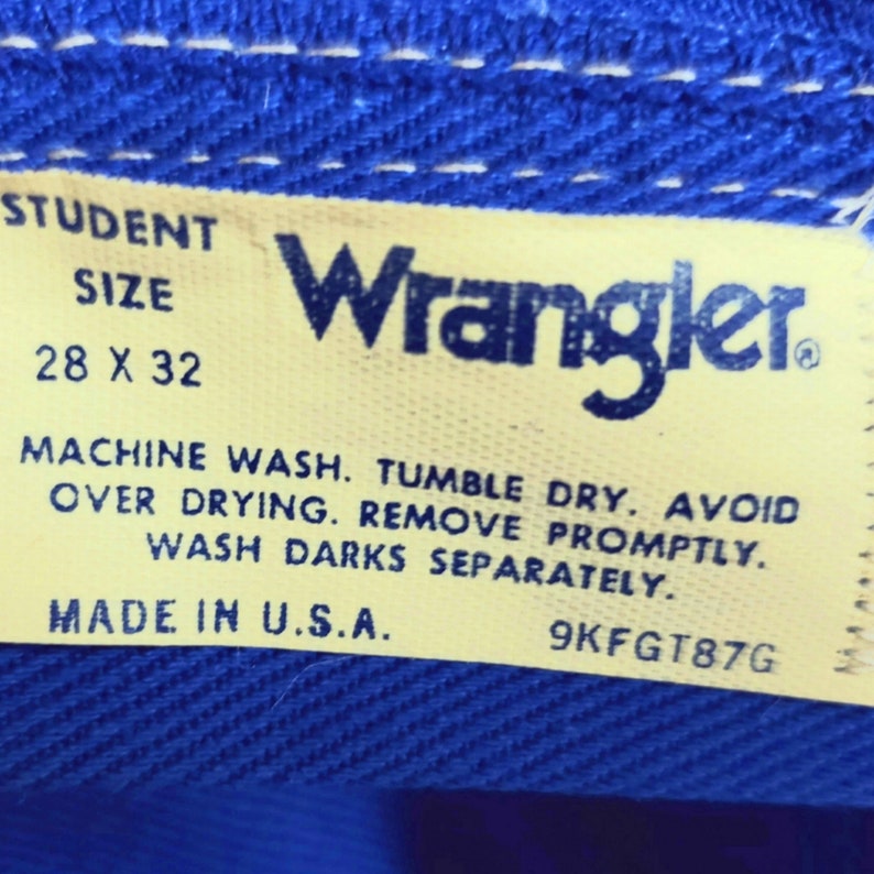 May include: A yellow Wrangler clothing care label with the text "STUDENT SIZE 28 X 32" and care instructions: "MACHINE WASH. TUMBLE DRY. AVOID OVER DRYING. REMOVE PROMPTLY. WASH DARKS SEPARATELY. MADE IN U.S.A. 9KFGT87G"