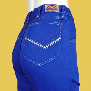 May include: Blue denim jeans with a back pocket featuring white stitching and a rainbow-coloured Wrangler logo.