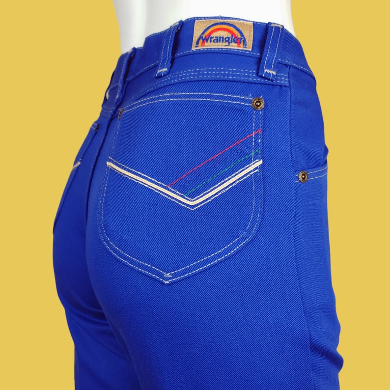 May include: Blue denim jeans with a back pocket featuring white stitching and a rainbow-coloured Wrangler logo.