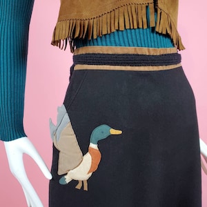 May include: A black skirt with a brown belt and a duck applique on the front pocket. The skirt has a fringe detail at the top.