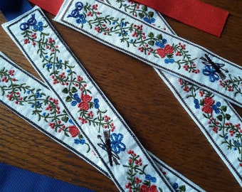 Puede incluir: Cinta blanca bordada con un diseño floral en rojo, azul y verde. La cinta tiene un borde negro y se combina con cintas rojas y azules. Las cintas están dispuestas sobre una superficie de madera.