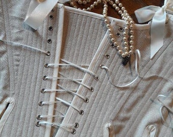 May include: A white cotton corset with white satin ribbon ties and a pearl necklace. The corset has a lace-up back with metal eyelets.