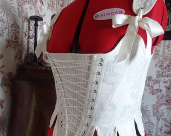 May include: A white corset with a floral pattern and a satin ribbon detail. The corset is laced up the front and has a scalloped hem. The corset is on a red dress form with a "Singer" label.
