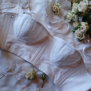 May include: A white strapless corset top featuring structured seams, silver-toned metal eyelets, and lace-up ribbon detailing. The garment is displayed alongside white fabric with lace trim and several small, pale cream-coloured roses.