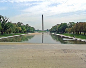 May include: A view of the Washington Monument in Washington, D.C. The monument is reflected in the still water of the reflecting pool. Trees line the sides of the pool, and people are walking along the paths.