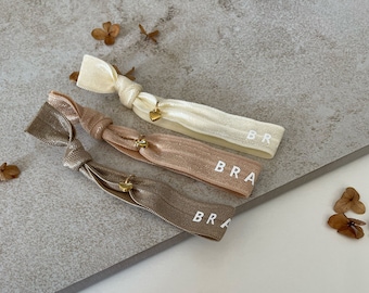 May include: Three hair ties with gold heart charms. The hair ties are in shades of beige, light brown, and cream. Each hair tie has the word "BRA" printed on it.