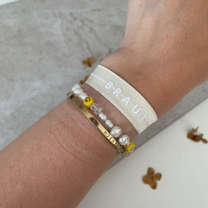 May include: A white elastic wristband with the word "BRAUT" printed on it. The wristband is worn on a person's wrist along with two bracelets. One bracelet is made of white pearls and yellow smiley face beads. The other bracelet is a gold chain with the word "FREUNDSCHAFT" engraved on it.