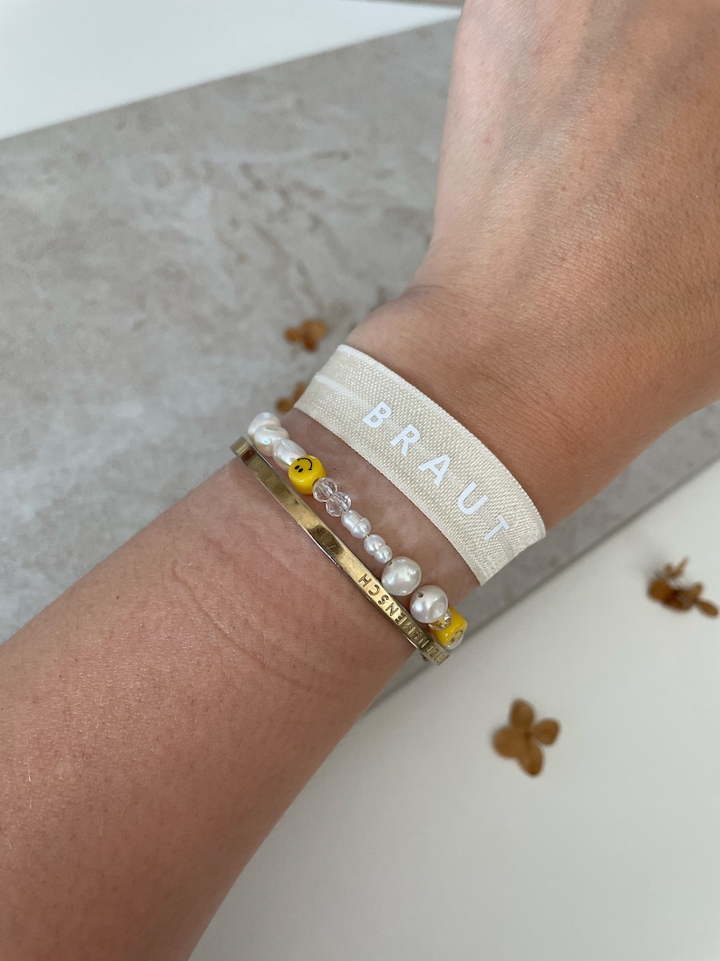 May include: A white elastic wristband with the word "BRAUT" printed on it. The wristband is worn on a person's wrist along with two bracelets. One bracelet is made of white pearls and yellow smiley face beads. The other bracelet is a gold chain with the word "FREUNDSCHAFT" engraved on it.