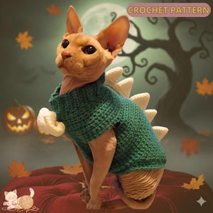 Puede incluir: Un gato sin pelo con un suéter de dinosaurio verde tejido con ganchillo y púas color crema. El gato está sentado en un cojín rojo con un fondo temático de Halloween. El texto "CROCHET PATTERN" es visible.
