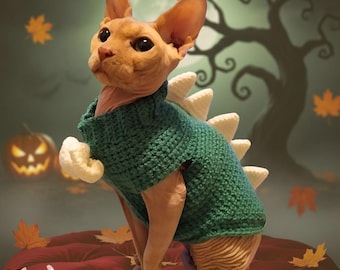Può includere: Un gatto nudo indossa un maglione a forma di dinosauro verde all'uncinetto con punte color crema. Il gatto è seduto su un cuscino rosso con uno sfondo a tema Halloween. Il testo "CROCHET PATTERN" è visibile.
