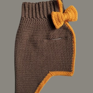 May include: A brown crocheted dog sweater with a yellow crocheted bow on the shoulder. The sweater has a yellow trim around the edges.