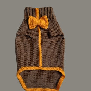 May include: A brown crocheted dog sweater with a yellow bow tie. The sweater has a yellow trim around the edges.