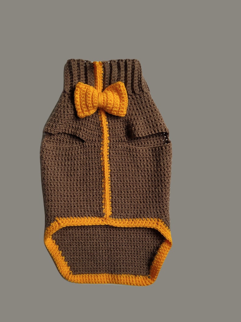 May include: A brown crocheted dog sweater with a yellow bow tie. The sweater has a yellow trim around the edges.