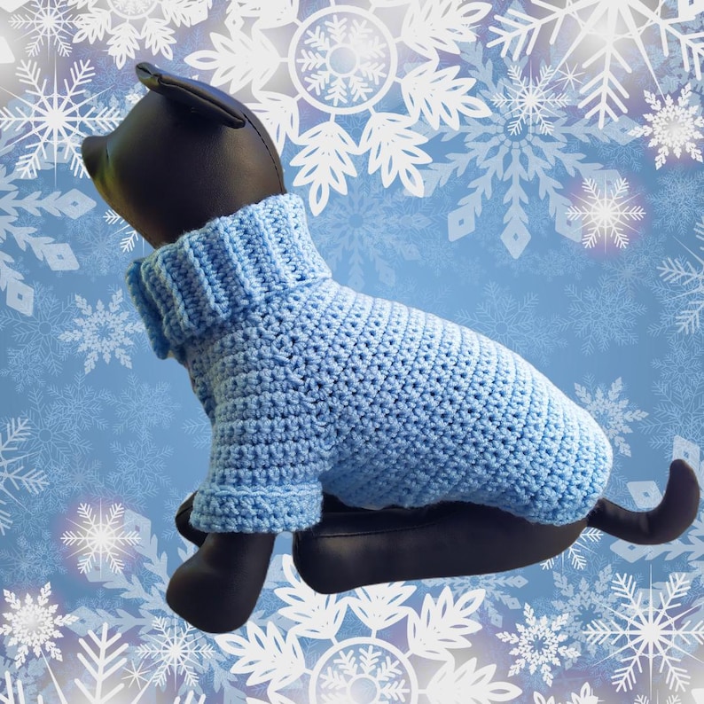 May include: A light blue crocheted dog sweater with a turtleneck. The sweater is on a mannequin dog against a blue background with white snowflakes.