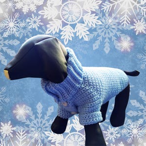 May include: A light blue knitted dog sweater with a button closure. The sweater is on a mannequin dog against a blue background with white snowflakes.