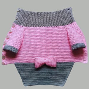 May include: A pink and grey crocheted sweater with a bow on the front. The sweater has buttons down the front and is made with a soft yarn.