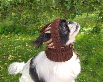 May include: A small white and black dog wearing a brown and beige knitted hat with a hood. The dog is looking up and has its mouth slightly open.