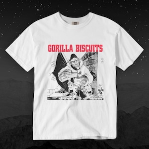 Pode incluir: Camiseta branca com o texto "GORILLA BISCUITS" em letras vermelhas sobre um gráfico em preto e branco de um gorila com capuz. O gorila está quebrando uma parede. A camiseta tem gola redonda.
