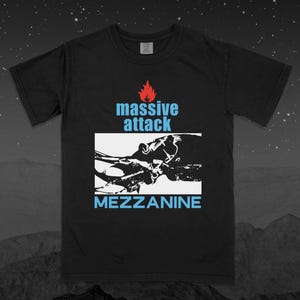 Puede incluir: Una camiseta negra de manga corta con un estampado gráfico que incluye un icono de llama roja sobre el texto azul claro "massive attack". Debajo, un recuadro blanco contiene la silueta negra de un escarabajo y el texto "MEZZANINE".