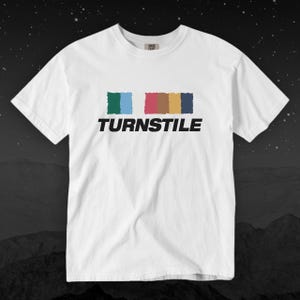 Könnte beinhalten: Weißes T-Shirt mit dem Wort "TURNSTILE" in schwarzen Blockbuchstaben. Über dem Wort befinden sich vier farbige Rechtecke: grün, blau, rosa, gold und marineblau. Das T-Shirt hat einen Rundhalsausschnitt und kurze Ärmel.
