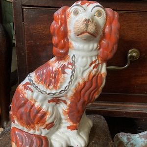 May include: A ceramic dog figurine featuring a white base with reddish-brown patches on the ears and body. The dog has painted facial features, including wide eyes and a snout, and a painted chain design draped across its chest and back.