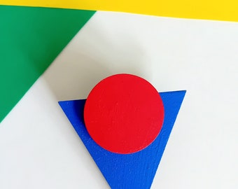 Könnte beinhalten: Ein geometrisches Holzkunstwerk mit einem roten Kreis auf einem blauen Dreieck. Das Kunstwerk befindet sich vor einem Hintergrund aus gelben, grünen und weißen geometrischen Formen, wodurch eine lebendige, abstrakte Komposition entsteht.